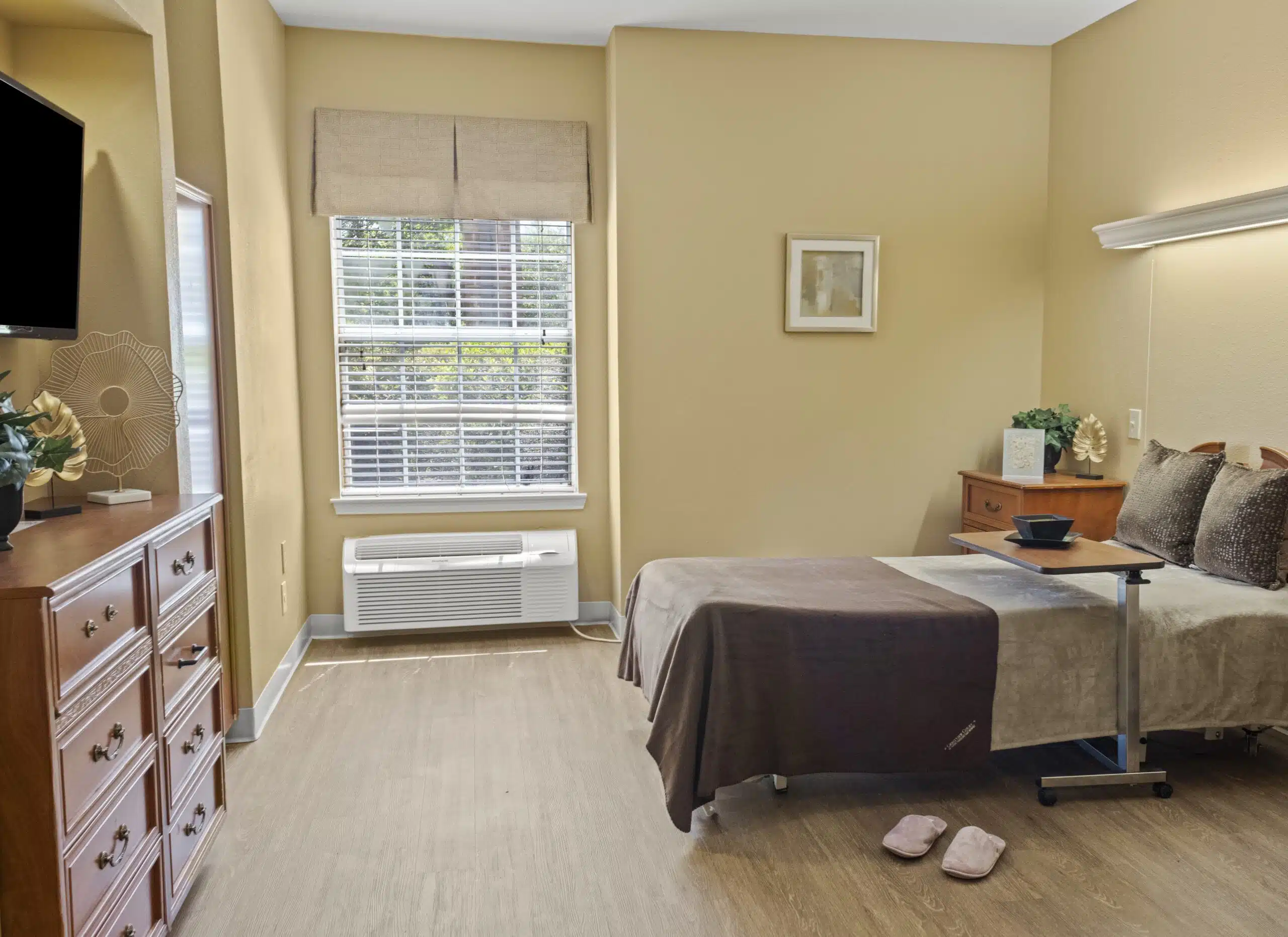 Bedroom photo with twin bed on right with a bedside roller table. Straight ahead is a window with heater/AC unit below. A chest of drawers is on the left. Tan paint on the walls