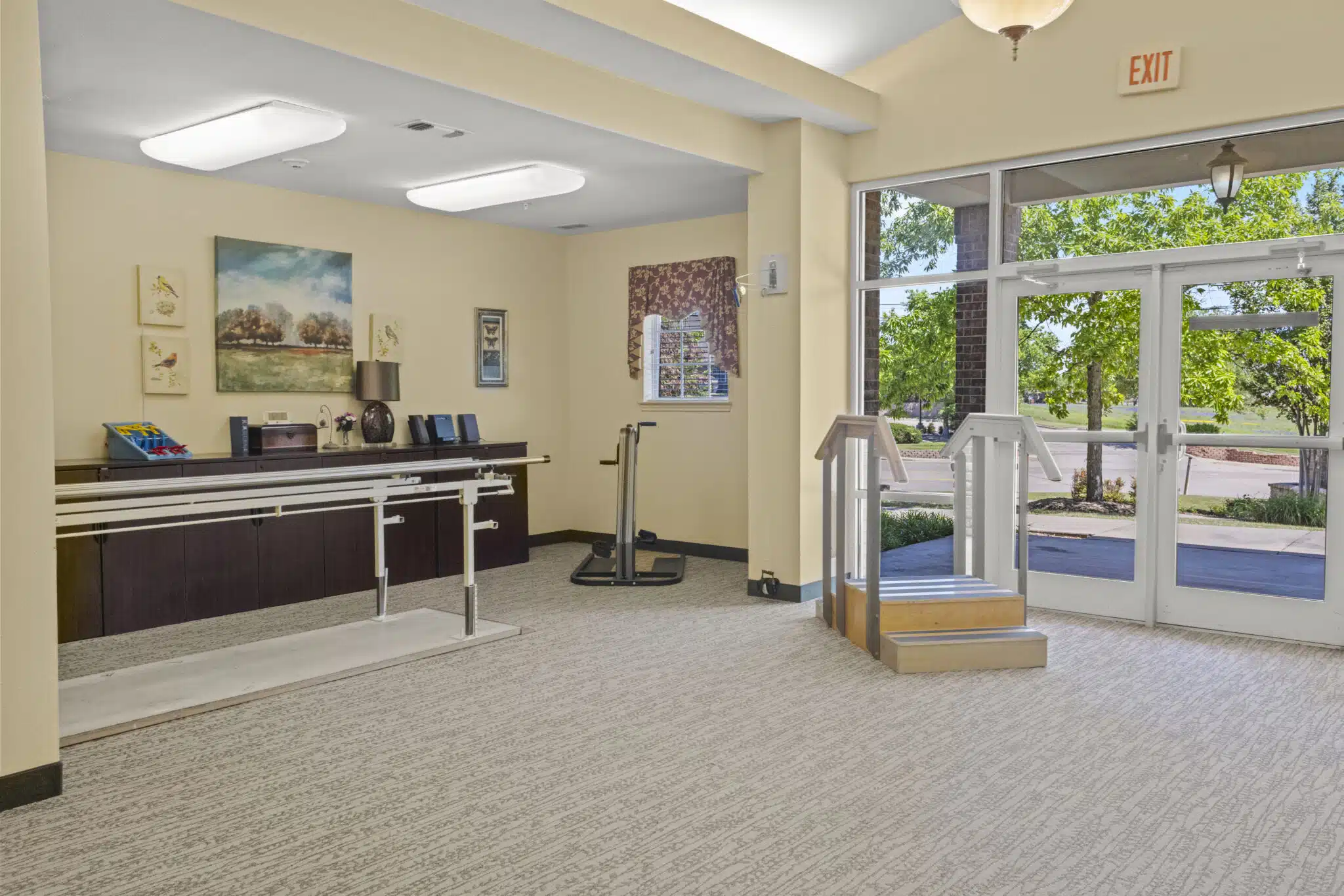 Another photo of the rehab gym with a stair climber, exercise bike, and parallel bars for rehabilitating residents who have difficulty walking. This gym faces an exit with a view of the outside.